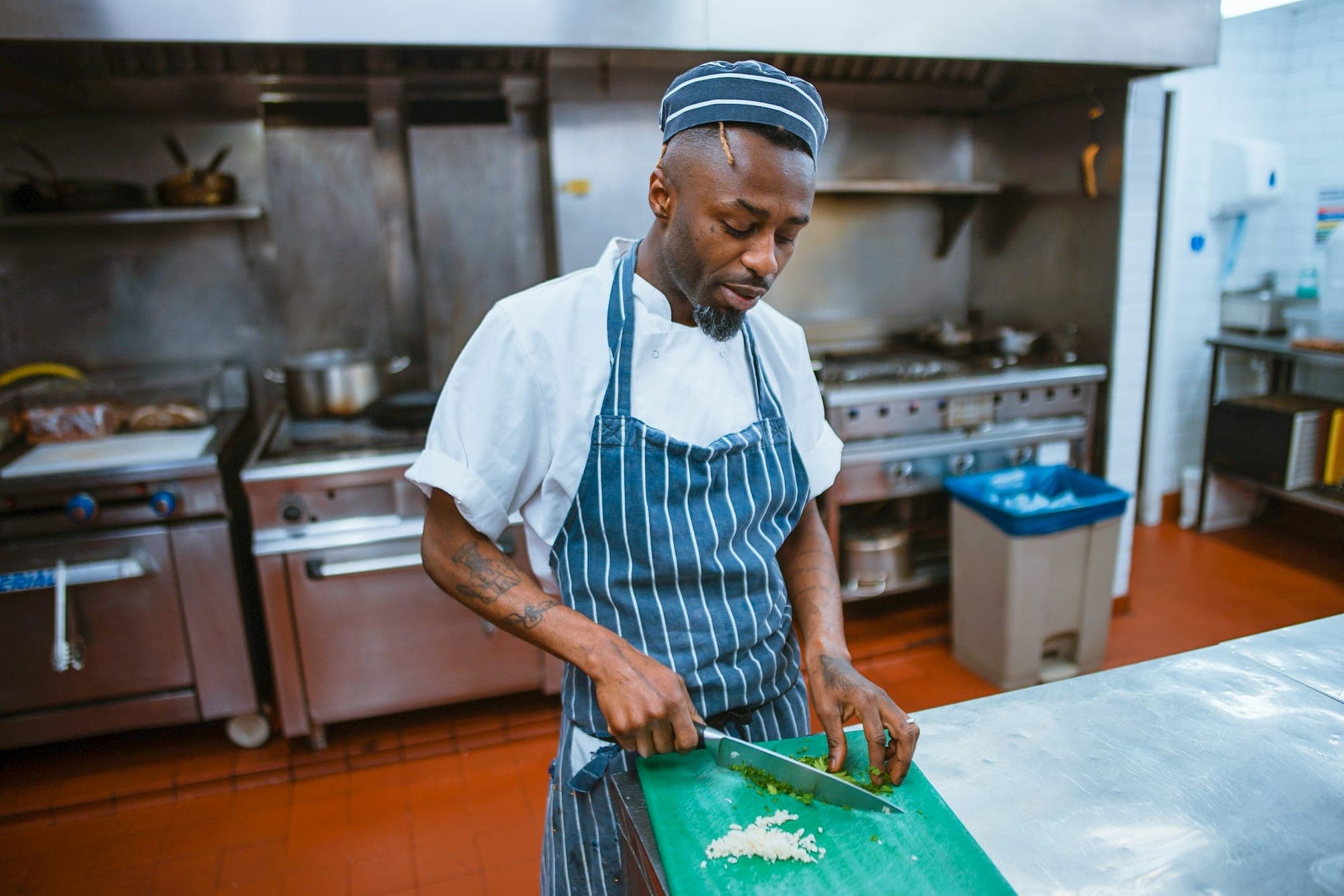 A chef in a striped apron chopping herbs on a green board in a commercial kitchen